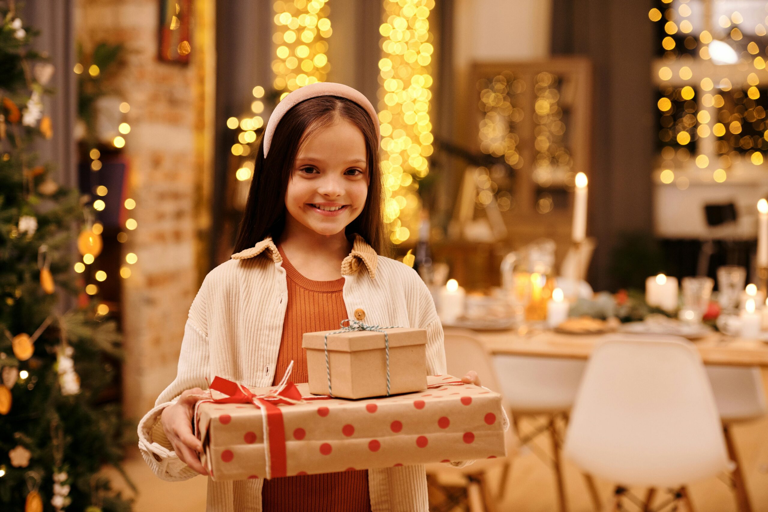 A girl smiles while holding a gift box in front of a beautifully decorated Christmas tree.