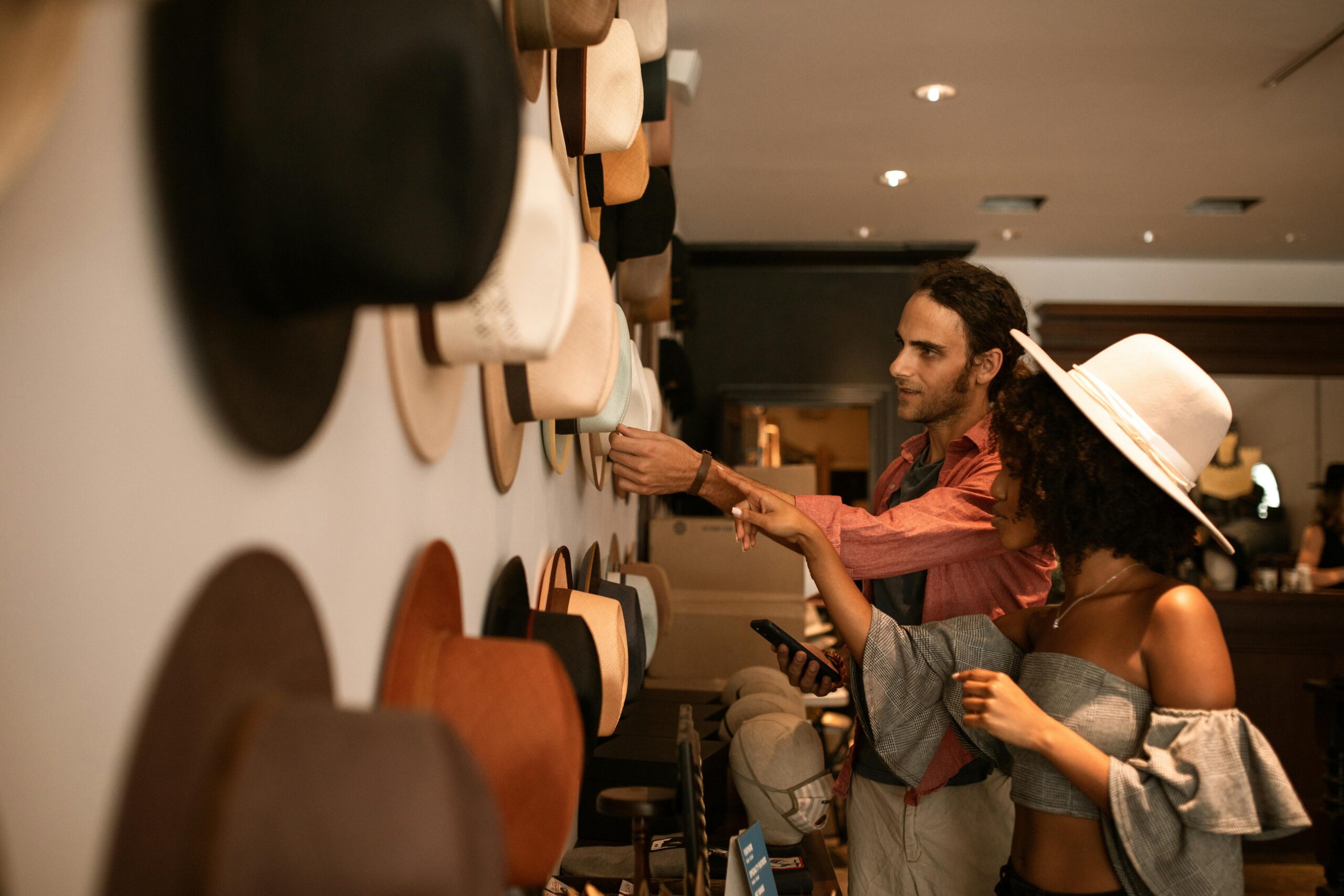 A man and woman examine various hats displayed on a wall in a store.