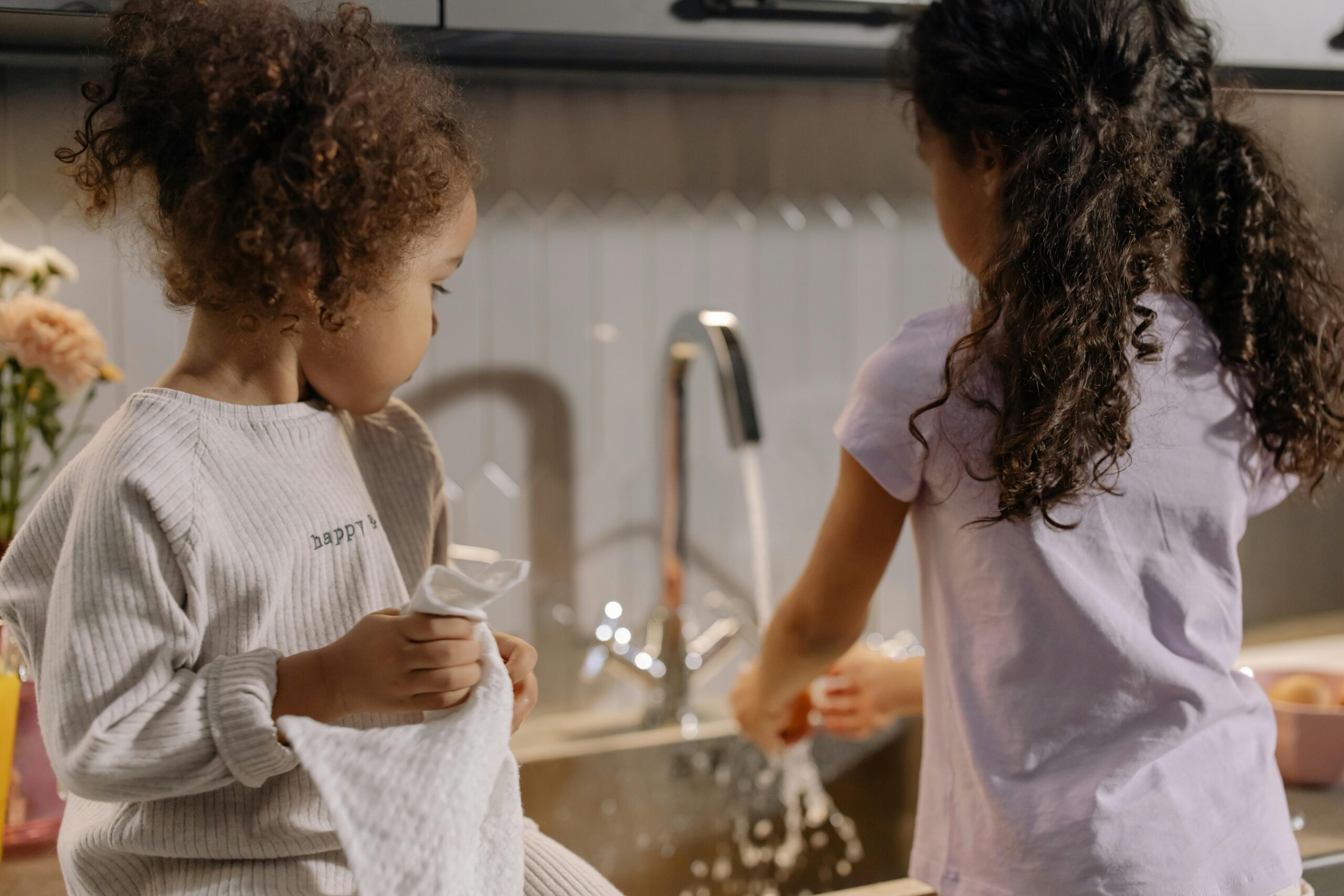 Two young girls happily washing dishes together in a kitchen sink, surrounded by soap bubbles and kitchen utensils.