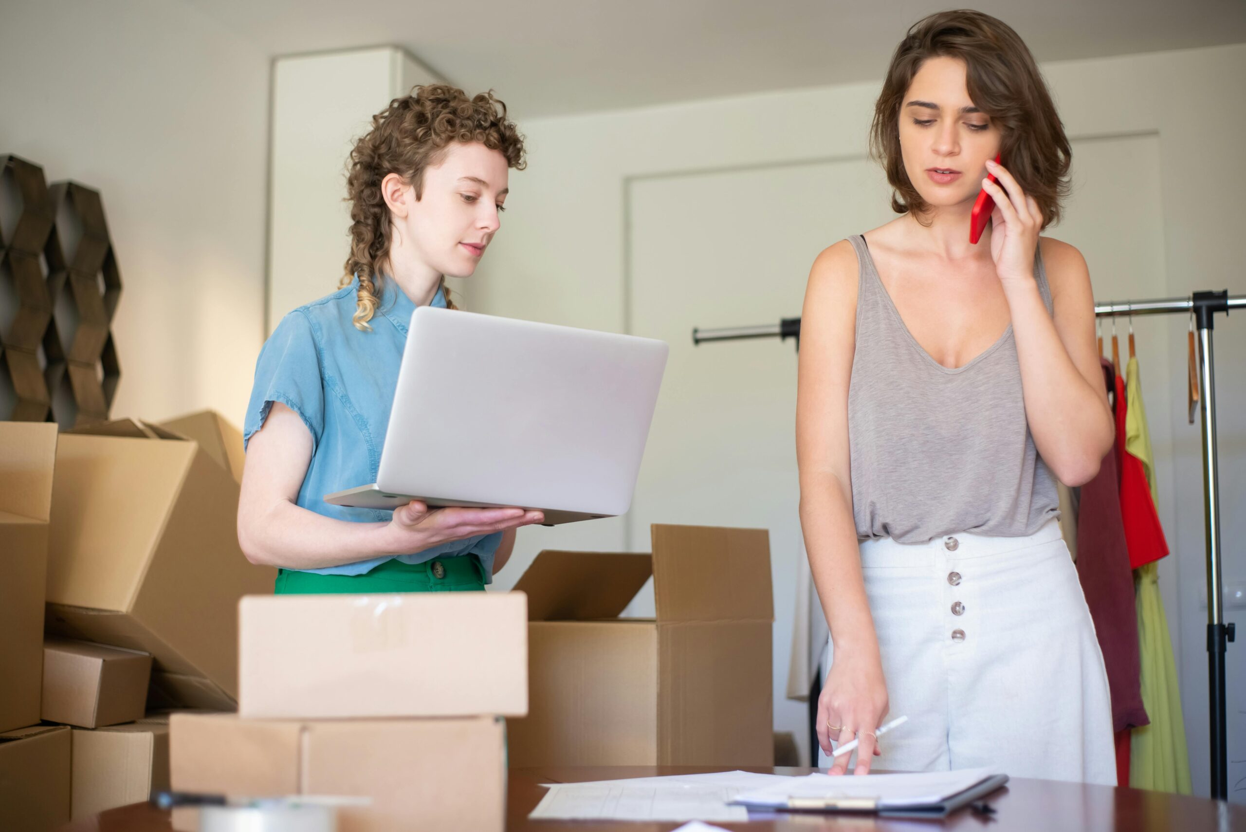 Two women stand beside boxes and a laptop, engaged in discussion or planning.