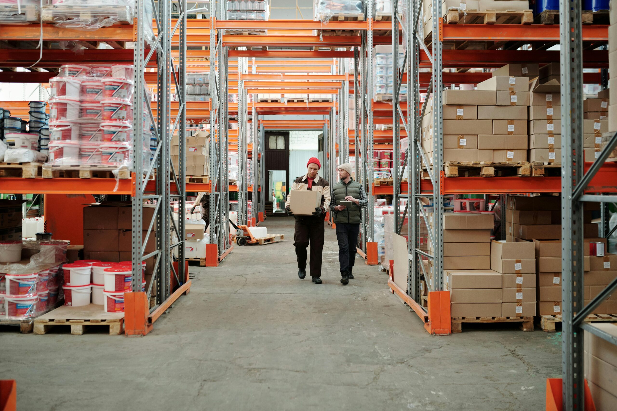 Two individuals walking through a warehouse filled with stacked boxes, navigating the aisles between them.