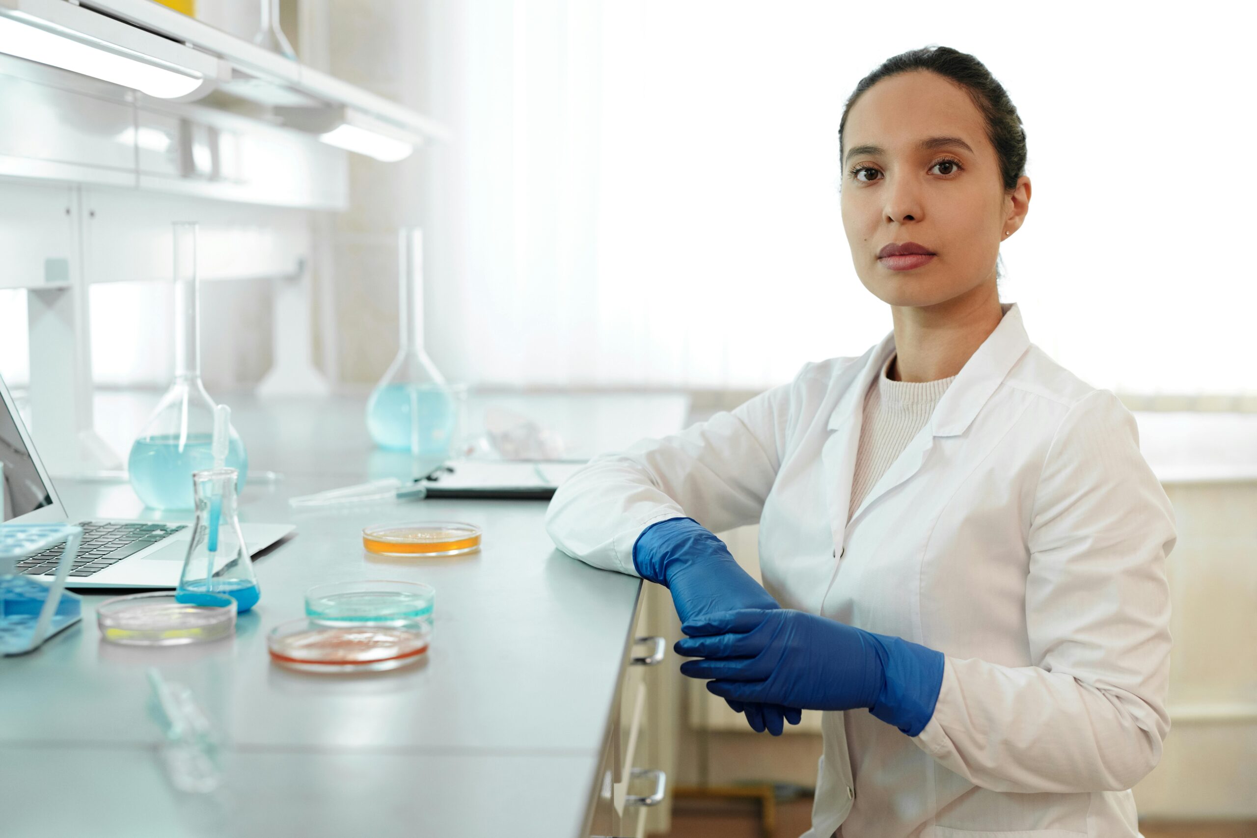 A woman in a lab coat and gloves sits at a desk, focused on her work in a laboratory setting.