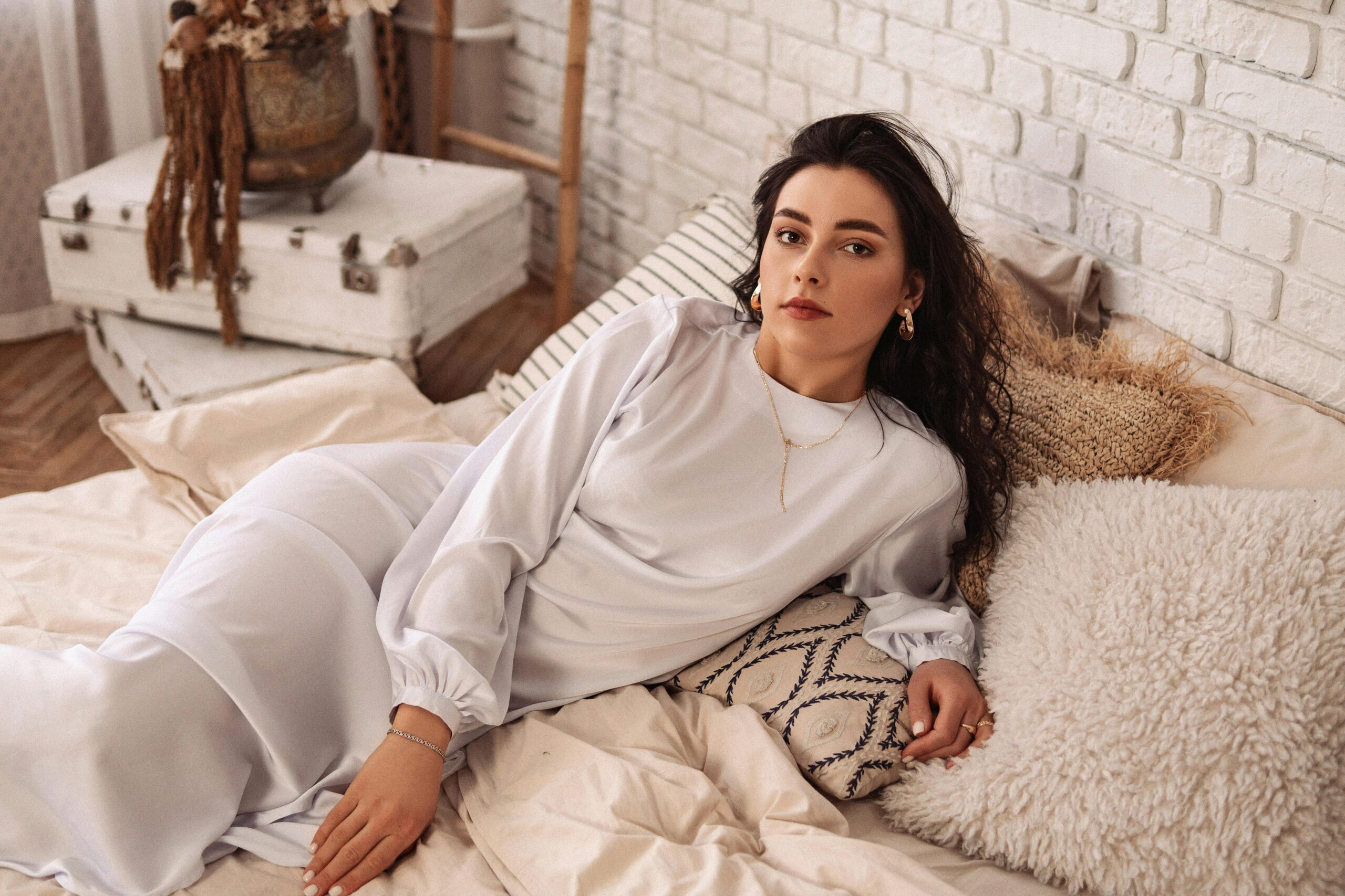 A woman in a white dress reclines gracefully on a neatly made bed, surrounded by soft pillows and natural light.